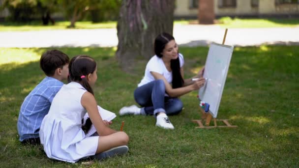 母亲用白板在新鲜空气中给她的两个孩子上一课 男孩和女孩听母亲或老师坐在屋外的绿草上 家庭教育概念 — 图库视频影像