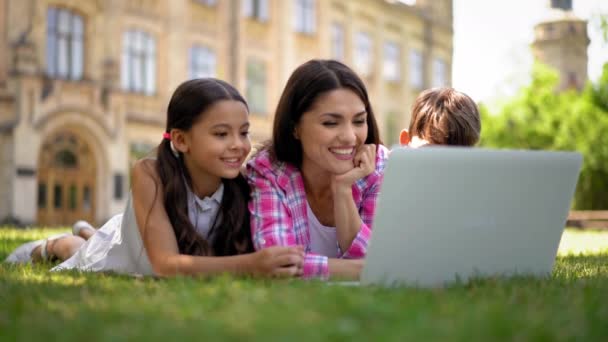 公園の芝生の上に寝そべっているノートパソコンで 母親と一緒にトレーニング漫画を見ている子供たち 家庭教育として屋外レッスンを受けている息子と娘を持つ母親 — ストック動画
