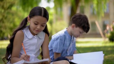 Okulun yanındaki yeşil çimlerde oturan ve resim çizen iki öğrencinin ön görüntüsü. Çocuklar açık kapılar sanat dersi alıyor. At kuyruklu kız ve sınıf arkadaşı açık havada defter çiziyor..