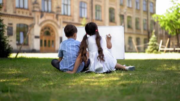 屋外に座っている2人の生徒の背面ビューと古代の学校の背景に白いボードを使用して勉強します 2人の子供の男の子と女の子は緑の草の上に座って新鮮な空気について勉強する — ストック動画