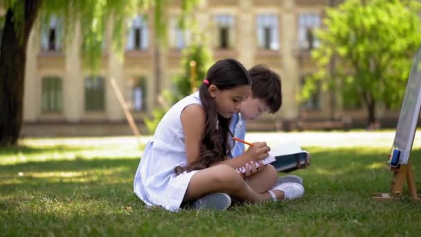 男の子と女の子 学校を背景に草の上に座って ノートに屋外の書き込みや絵の性質を勉強する — ストック動画