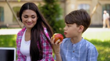 Anne gülümsüyor ya da gülüyor. Oğlunun çerez olarak elma yemesini izliyor. Parktaki yeşil çimlerin üzerinde oturuyor..