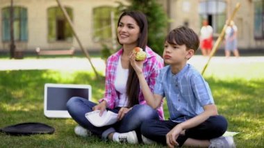 Anne ve oğlu okuldan sonra temiz havada. Annesi uzaktan bakarken oğlu çim tezgahının yanında elma yiyor. İşle iletişim kurmak için kullandığı bir dizüstü bilgisayar ya da çizgi film izliyor..
