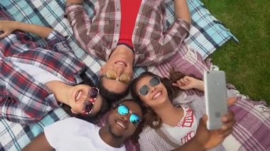Güzel genç insanlar dışarıda çimenlerde uzanmış akıllı telefonlarıyla selfie çekiyorlar..