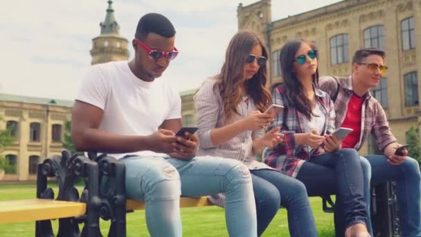 Amigos sentados en benchand mensajes de texto en teléfonos celulares — Vídeo de stock