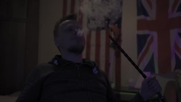 Homem na sala de luz escura fumando narguilé e refrigeração . — Vídeo de Stock