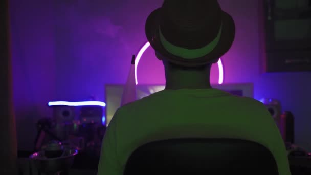 Homem usando chapéu na sala de luz de néon fumando narguilé . — Vídeo de Stock