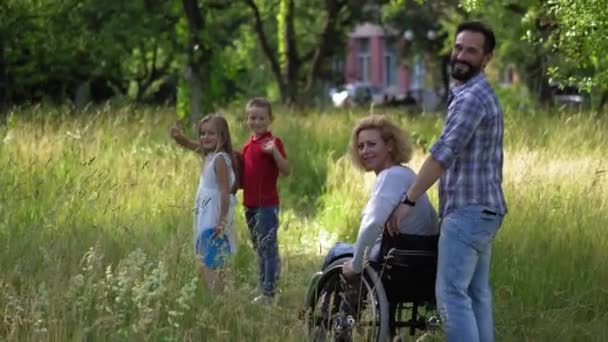母亲坐在轮椅上的家庭回头看过去，在镜头前挥挥手. — 图库视频影像
