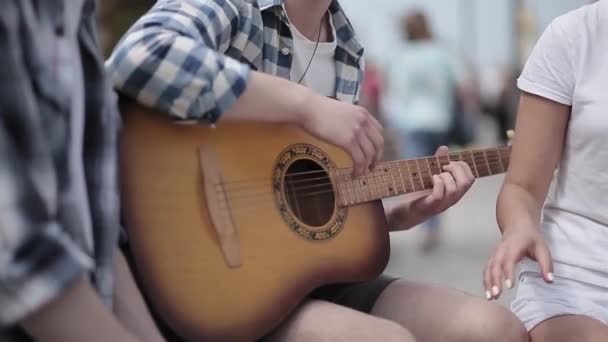 Młodzi ludzie siedzą w mieście na ulicy i grają na gitarze, a dwóch przyjaciół siedzi obok i śpiewa. Zbliżenie gitary w rękach faceta. Prores 422 — Wideo stockowe