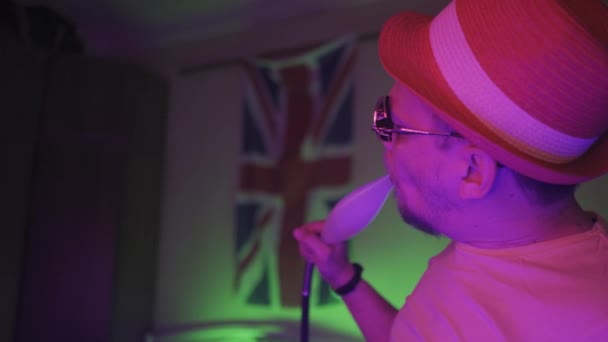 Rauchender Shisha-Hipster aus Großbritannien mit Sonnenbrille in dunklem, halb beleuchtetem Neonlicht-Raum sitzt liegend auf dem Stuhl und reitet seitlich mit der UK-Flagge an der Wand — Stockvideo