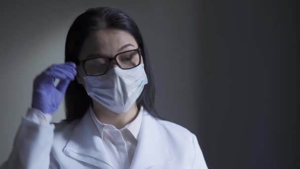 Уставшая микробиолог азиатка на рабочем месте снимает маску и очки и прикасается к лицу одноразовыми перчатками. Усталость ведет к неосмотрительности. Прорес 422 — стоковое видео