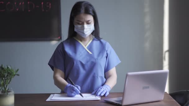 Sköterskan gör en skylt där det står att hon lyfter upp den framför sig själv med medicinsk uniform och föroreningsmask framför laptopen på bordet. Prores 422. — Stockvideo