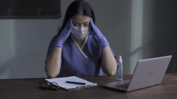 Ένα άτομο που κάθεται σε ένα γραφείο μπροστά από ένα φορητό υπολογιστή με ένα κουρασμένο πρόσωπο και κρατώντας το κεφάλι με καθαρισμένα αντιβακτηριακά υγρά γάντια. Πονοκέφαλος στην εργασία πόνους νοσοκόμα. Πρότυπα 422 — Αρχείο Βίντεο
