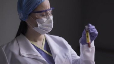 Bir mikrobiyolog üzerinde COVID 19 etiketi olan hastanın kanıyla test tüpünü kontrol eder. Kadın doktor, pandemik koronavirüs sırasında yorgun. Kapatın. Prores 422