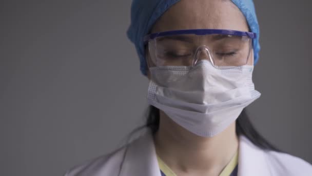 Enfermera mirando cuidadosamente a la cámara con un uniforme médico, una máscara protectora y una gorra y gafas de protección. Enfermera de etnia asiática durante una pandemia. Prores 422 — Vídeo de stock