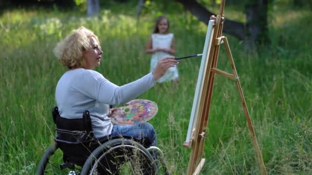 Художник у інвалідному візку прикрашає портрет маленької дівчинки. Молода модель втомилася і намагається рухатися. Художник з мольбертом у природі. Проходить 422 — стокове відео