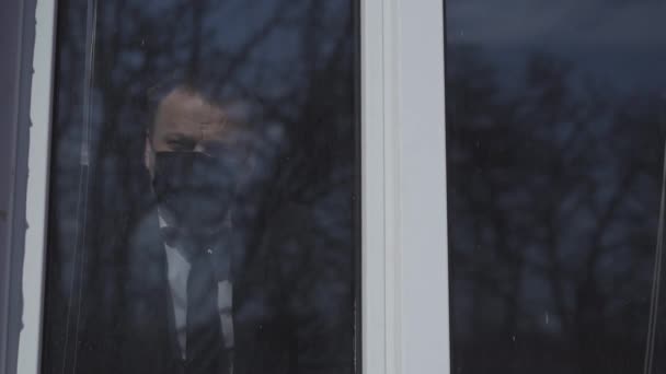 Άντρας με προστατευτική μάσκα και επαγγελματικό κοστούμι στέκεται στο παράθυρο και ψάχνει για το τι συμβαίνει έξω από το κτίριο. Εργασία κατά τη διάρκεια της περιόδου καραντίνας της πανδημίας coronavirus. Πρότυπα 422 — Αρχείο Βίντεο