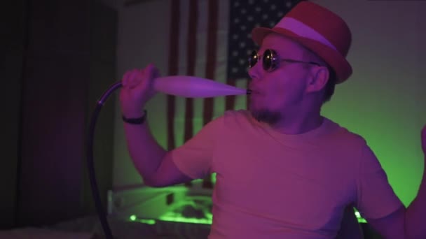 A fiatalember vízipipát szív az amerikai zászló hátterében, aktív zenét hallgat és táncol a neon félig megvilágított fényszobában. Gyümölcsfélék — Stock videók