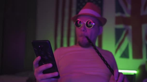 En man som sitter i ett mörkt rum med smartphone i händerna och bär solglasögon och hatt när han röker hookah. Hipster sitter i ett halvbelyst neonljus rum. Prores 422 — Stockvideo