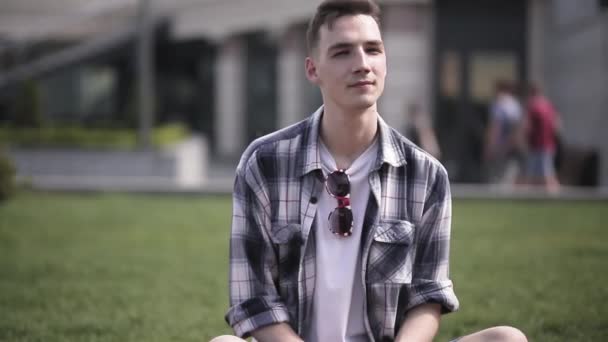 若い男が街の芝生の上に座っている友人を待っている。チェックしたシャツを着た男が、芝生の上に座っている街の背景にガールフレンドを待っている。第四百二十二条 — ストック動画
