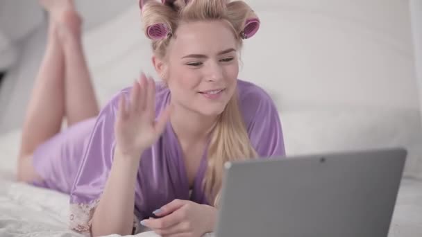 En tjej på självisolering sitter hemma och kommunicerar med vänner via videolänk medan hon ligger med en laptop på sängen. en flicka med lockigt hår kommunicerar med sin vän på Internet — Stockvideo