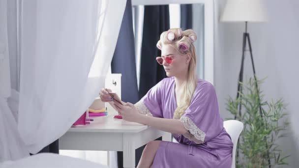 Die Blondine mit pinkfarbener Brille und Morgenmantel sitzt mit dem Handy in der Hand am Fenster und chattet mit jemandem oder surft in sozialen Netzwerken. Blond auf Selbstnachahmung. Prores 422 — Stockvideo