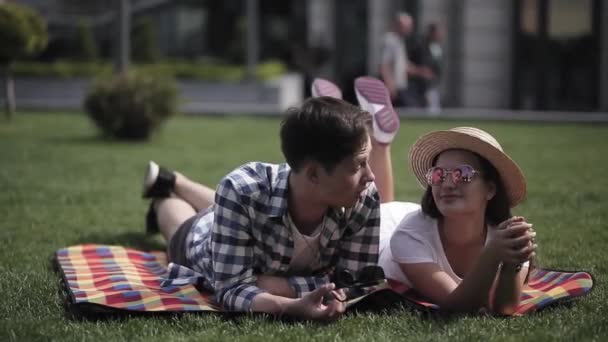 Young people are in love and spend time together lying on the lawn on a blanket with printed cubes. young man taking off his glasses looks at his girlfriend in a hat. Prores 422 — Stock Video