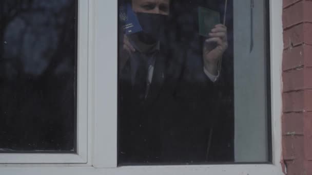 Hombre de negocios encerrado frente a una ventanaHombre de cuarto en una suite de negocios parado frente a una ventana que muestra pasaporte o documentos en las manos tratando de llamar la atención de las personas o los médicos de los CDC — Vídeo de stock