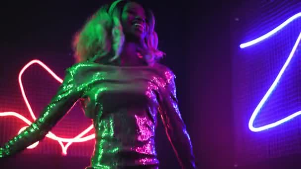 Chica afroamericana bailando feliz frente a una pared de luces de neón. Chica con un vestido de lentejuelas plateadas. Concepto de neón. Prores 422 — Vídeos de Stock