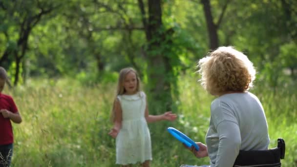 Mamma su una sedia a rotelle gioca con se stessa sulla linea gettando Frisbee ai bambini e papà è dietro di loro una sicurezza. Concetto familiare. Prore 422 — Video Stock