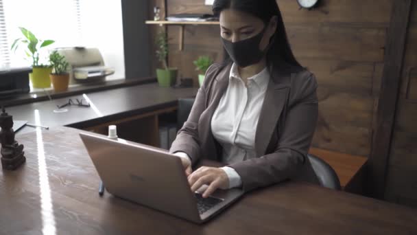 Mulher em máscara protetora trabalhando no escritório sentado em uma mesa usando laptop. Mulher asiática vestindo roupas de negócios trabalhando durante dias de quarentena em um escritório loft. Prores 422 — Vídeo de Stock