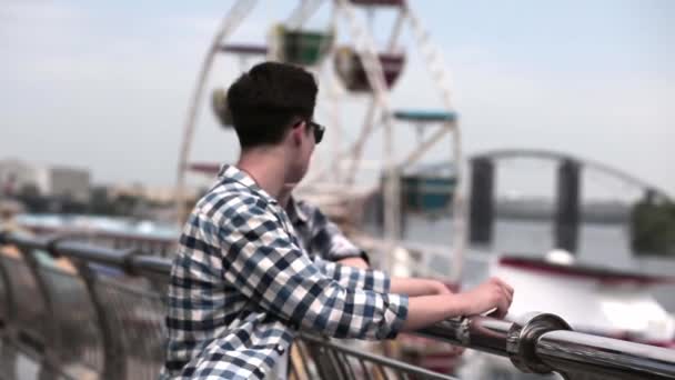 Dois amigos estão de pé perto de trilhos para uma área de entretenimento a pé no dique do rio com uma roda gigante. Dois caras de camisas xadrez ficar e olhar para a vista da cidade. Foco seletivo. Prores 422 — Vídeo de Stock