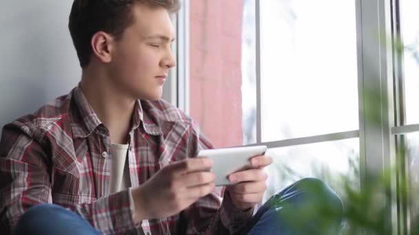 男は窓のそばの窓辺に座っている間、スマートフォンで遊んでいる。学生は、ソーシャルネットワークやビデオをレビューすることによって試験の準備から気を取られています.学生のコンセプト。第四百二十二条 — ストック動画