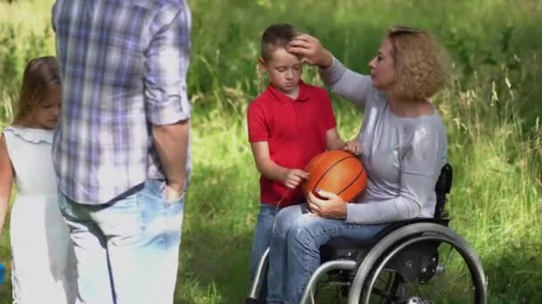 Mom in a wheelchair throws frisbee to his son and father plays with his daughter throwing a ball on nature in the park after a picnic. Family concept. Prores 422 — Stock Video