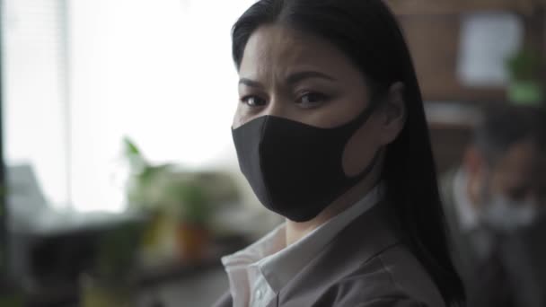 保護マスクをしたアジア系の実業家がカメラを見て窓の外を眺めている。私はコロナウイルスのパンデミックの間にオフィスで働いています。隔離の概念で働く。第四百二十二条 — ストック動画