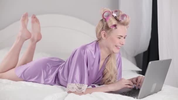 Блондинка з ноутбуком і кучеряве волосся у відеодзвінку з батьками з-за кордону. Дівчина з карантином спілкується з друзями або дівчиною, коли лежить на ліжку. Залишайтеся вдома концепції. Проходить 422 — стокове відео