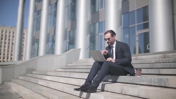 ビジネススーツの男は、ビジネスセンターの背景にあるステップに座ってラップトップで動作します。ビジネスコンセプト。第四百二十二条 — ストック動画