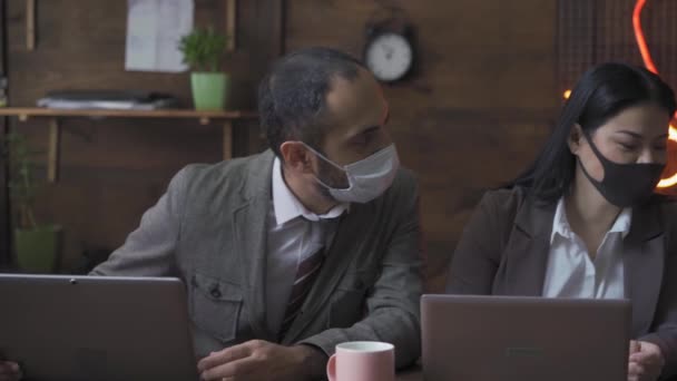 Les pigistes masqués protecteurs travaillent au bureau pendant la période de quarantaine. Les jeunes en tenue de travail discutent des questions de travail tout en étant assis à une table avec des ordinateurs portables. Concept d'entreprise — Video