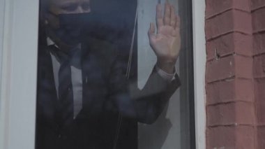 Takım elbiseli ve koruyucu maskeli bir adam pencerenin önünde kameraya el sallıyor. Karantina salgını sırasında kendini izole eden iş adamı. Prores 422