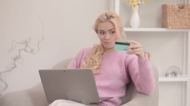 Blond girl holding a credit card with laptop doing online shopping or payments. Young blond woman buying a dress online. Fashion model buying food in online shop during self isolation. Prores 422 — Stock Video