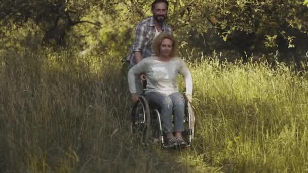 Les enfants apportent des fleurs sauvages à une mère en fauteuil roulant. Père aider mère dans un fauteuil roulant pour obtenir à travers l'herbe haute dans le parc ou arrière-cour. Concept de famille. Prores 422 — Video