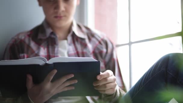 Egy diák könyvet olvas, miközben az ablakpárkányon ül. Egy fiatalember a szabadidejét karanténban töltött irodalom olvasásával tölti az elszigeteltség időszakában. Maradj otthon! Gyümölcsfélék — Stock videók
