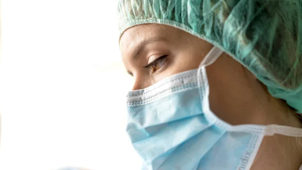 Endişeli, koruyucu kıyafetli bir kadın doktor, hastane penceresinden bakıyor.