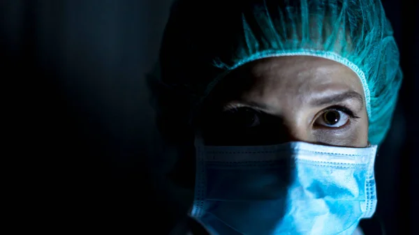 Portrait of young female surgeon, wearing mask and a surgical mask, in front of black background