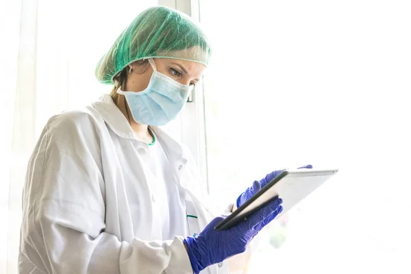 Hastanede dikilirken dijital tablet kullanan tanınmayan bir kadın hemşirenin görüntüsü. Bir maske tak