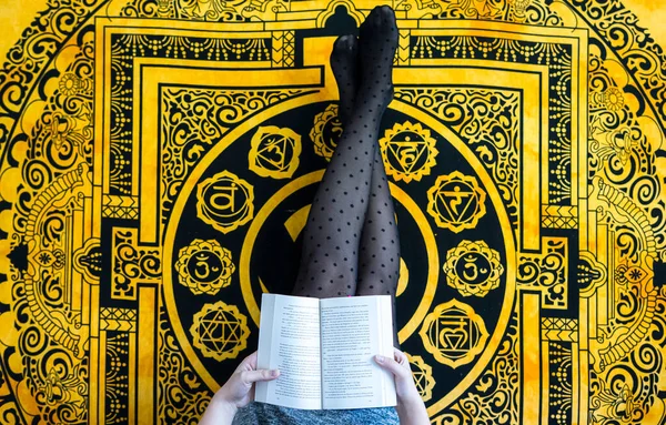 Yatağa uzanmış, bacakları kalkmış ve sarı arka planda kitap okuyan kadın..