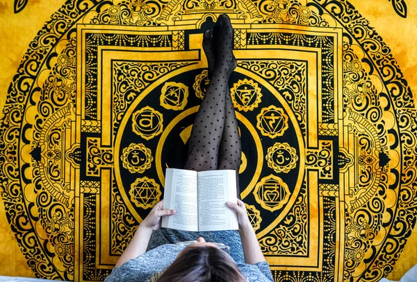 Yatağa uzanmış, bacakları kalkmış ve sarı arka planda kitap okuyan kadın..
