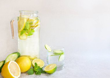 Uzun bir sürahide limonata ve gri arka planda cam ferahlatıcı. Limon, limon, salatalık ve nanenin yanında. Yatay konum, metin için boşluk