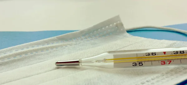 Civa termometresi tek kullanımlık tıbbi bir maskenin üzerinde. Termometre yüksek ateşi gösteriyor. Tıbbi maske beyazdır. Virüsten koruma. Mavi arka plan. Coronavirus. Uzayı kopyala Pankart
