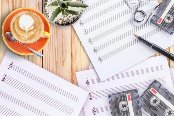 Hudba na notovém papíře, kaktus, plnicí pero, kazeta s páskou a káva latte na dřevěný stůl — Stock fotografie
